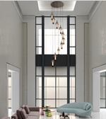 A modern multi-light linear chandelier with copper-toned glass cylinders hangs in a tall, contemporary living room with a high ceiling, black-framed windows, a light blue lounge chair, and a sofa.