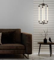 A modern multi-light pendant chandelier with four vertical frosted glass tubes and a dark metal frame, hanging in a contemporary living room next to a brown sofa and a striped window valance.