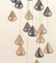 A modern multi-light chandelier featuring multiple teardrop-shaped glass pendant shades in smoked silver and warm champagne beige, suspended by thin cables against a solid off-white background.