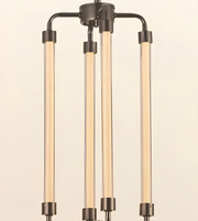 A modern three-light linear chandelier with a dark gunmetal finish and warm glowing frosted glass tubes, shown against a solid cream background.