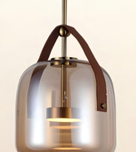 A modern pendant light with a rounded, translucent smoky glass shade, a vertical brass rod, a brown leather strap, and a warm glowing light inside, shown against a plain off-white background.