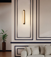 A modern vertical wall sconce with a warm glowing cylindrical light tube and brass-toned end caps, mounted on a beige wall with dark decorative panel lines above a light sofa.