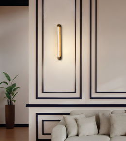 A modern vertical wall sconce with a warm glowing cylindrical light tube and brass-toned end caps, mounted on a beige wall with dark decorative panel lines above a light sofa.