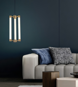 A modern three-light linear pendant chandelier with vertical frosted glass tubes and brushed brass hardware, hanging in a contemporary living room above a light gray sofa with blue and black patterned pillows against a dark navy wall.