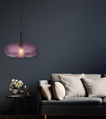 A modern pendant light with a textured, woven purple shade hangs over a dark gray sofa with beige pillows in a dimly lit living room.