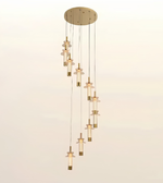 A modern multi-light linear pendant chandelier with a circular brushed brass ceiling canopy, slim hanging cords, and multiple cascading glass shades with brass accents, shown against a plain light background.