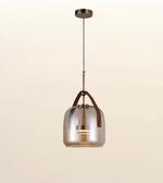 A modern pendant light with a clear glass bell-shaped shade, a brass-toned metal rod and canopy, and brown leather carrying handles, shown against a plain light beige background.
