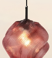 A close-up of a hanging pendant light with a ribbed, translucent glass shade in a dusty rose or mauve pink color, illuminated from within, against a plain light beige background.