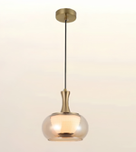 A modern pendant light with a warm brass-finished metal canopy, cord, and neck, paired with a large clear glass shade that glows with a warm yellow light, shown against a plain off-white background.