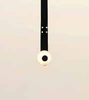 A minimalist pendant light with a black cord and a spherical bulb that glows warm white, suspended against a plain light beige background.