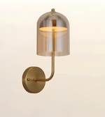 A modern wall-mounted sconce with a warm brass-finished metal body and a translucent smoky amber glass dome shade, shown against a plain light beige background.