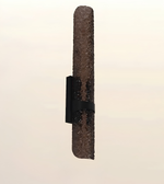 A modern wall-mounted light fixture with a textured, rusted brown cylindrical column and a black mounting bracket, shown against a plain off-white background.