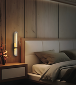 A modern wall-mounted vertical LED light fixture with a gold accent, illuminated and mounted on a light wood-paneled wall next to a bed with beige bedding.
