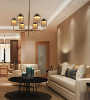 A modern living room interior featuring a multi-light chandelier with six glass bell shades and warm metallic arms, hanging above a black round coffee table. The room has beige walls, a cream sofa with patterned pillows, and a matching armchair.