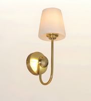 A modern wall sconce light fixture with a warm brass/gold metal frame and a tapered frosted white glass shade, shown illuminated against a plain off-white background.