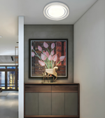 A modern ceiling light fixture with concentric circular rings, emitting a bright white glow, mounted on a white ceiling in a contemporary interior hallway.