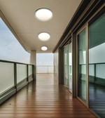 Modern outdoor balcony with a warm brown wooden deck, frosted glass railings, large sliding glass doors, and three round flush-mount ceiling lights.