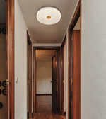A modern circular ceiling light fixture with a textured white glass outer ring and a small gold center, installed in a hallway with dark wood doors and flooring.