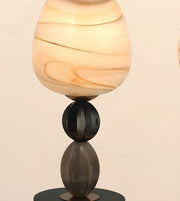 A table lamp with a rounded, teardrop-shaped shade made of marbled cream and tan glass, supported by a dark metallic base with reflective dark gray and black elements.