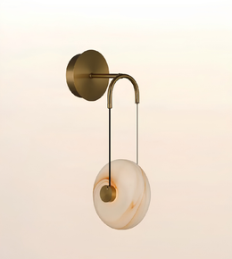A modern wall-mounted light fixture with a round white and beige marbled glass shade, suspended by a curved brass-toned metal arm from a circular wall plate, shown against a soft warm gradient background.