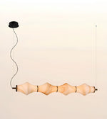 A modern multi-light linear pendant chandelier with five organic, wavy ribbed glass shades in a warm beige tone, suspended by a black cord from a black ceiling canopy against a light beige background.