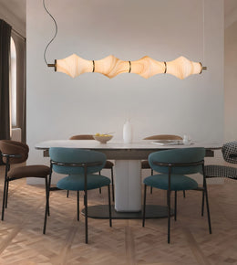 A modern dining room scene featuring a long horizontal pendant light with a wavy, segmented beige shade hanging above a white oval dining table with teal upholstered chairs.