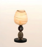 A modern table lamp with a rounded, teardrop-shaped shade made of marbled amber and cream glass, supported by a dark brown glass stem and a round black base, set against a soft beige gradient background.