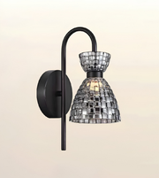 A modern wall sconce light fixture with a matte black curved arm and circular wall mount, featuring a clear glass shade made of reflective mirrored tiles that reflect warm light.