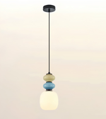A modern pendant light fixture with a black metal rod and canopy, featuring stacked frosted glass shades in cream, light blue, and white, glowing against a soft beige gradient background.