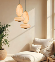 Two modern pendant light fixtures with organic-shaped cream and beige marbled glass shades hang in a softly lit room above a low cushioned daybed. A potted plant and a small wooden side table are visible in the background.