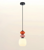 A modern pendant light fixture with a black metal rod, a round black ceiling canopy, and three stacked glass beads in orange, pink, and white, with the bottom shade glowing warm white.
