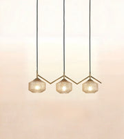 Three modern pendant lights with faceted glass shades and gold-toned geometric metal frames, suspended by black cords against a warm off-white wall.