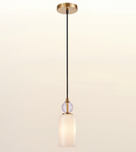 A modern single-light pendant lamp with a brushed gold ceiling canopy and trim, a black cord, a clear faceted glass sphere accent, and a frosted white glass cylindrical shade, shown against a warm off-white gradient background.