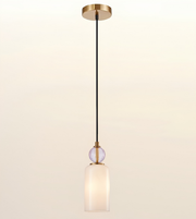 A modern single-light pendant lamp with a brushed gold ceiling canopy and trim, a black cord, a clear faceted glass sphere accent, and a frosted white glass cylindrical shade, shown against a warm off-white gradient background.
