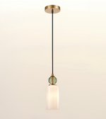 A modern single-light pendant lamp with a black cord, a round green glass bead accent, and a frosted white cylindrical shade, suspended from a brushed brass ceiling canopy against a warm off-white background.