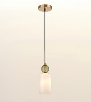 A modern single-light pendant lamp with a black cord, a round green glass bead accent, and a frosted white cylindrical shade, suspended from a brushed brass ceiling canopy against a warm off-white background.