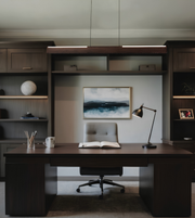 A modern home office setup featuring a dark wood desk and matching built-in shelving, with a gray tufted office chair in front of a framed landscape painting. A black desk lamp sits on the right side of the desk.
