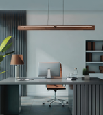 A modern minimalist office setup featuring a long rectangular wooden pendant light hanging above a gray desk with a computer, a brown leather swivel chair, and a matching table lamp.