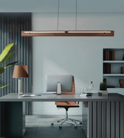 A modern minimalist office setup featuring a long rectangular wooden pendant light hanging above a gray desk with a computer, a brown leather swivel chair, and a matching table lamp.