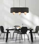 A modern dining room scene with a black rectangular dining table and four black chairs under a large black cylindrical pendant light with three glowing bulbs. The room has a minimalist white wall with soft shadows and a light gray floor.