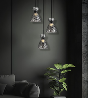 Three modern pendant lights with textured, mosaic-like silver shades hanging in a row against a dark charcoal wall, with a green potted plant and part of a dark sofa visible in the foreground.