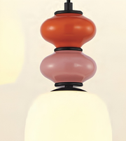 A close-up of a modern pendant light with stacked, rounded glass shades in terracotta orange and dusty pink, separated by black bands, with a glowing warm white glass shade below.