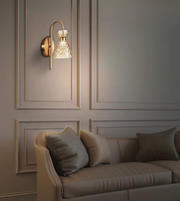 A modern wall sconce with a gold-finished curved arm and a textured, faceted clear glass shade, mounted on a beige paneled wall above a matching beige sofa with textured pillows.