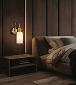 A modern wall-mounted sconce with a curved brushed brass arm, a round wooden finial, and a frosted white cylindrical glass shade, shown lit in a bedroom setting next to a bed.