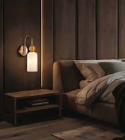 A modern wall-mounted sconce with a curved brushed brass arm, a round wooden finial, and a frosted white cylindrical glass shade, shown lit in a bedroom setting next to a bed.
