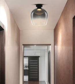 A modern semi-flush mount ceiling light with a clear glass dome shade and black metal hardware, shown installed in a hallway with warm ambient lighting.