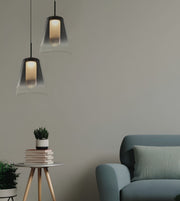 Two modern pendant lights with dark metal frames and clear glass shades hang in a minimalist room setting next to a blue-gray armchair and a small side table with books and a plant.