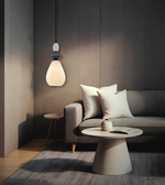 A modern pendant light with a glowing frosted glass shade hangs above a gray sofa in a minimalist living room setting with a round coffee table and neutral decor.