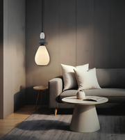 A modern pendant light with a glowing frosted glass shade hangs above a gray sofa in a minimalist living room setting with a round coffee table and neutral decor.