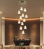 A modern multi-light pendant chandelier with 12 tapered white glass shades hanging in a staggered vertical arrangement from a circular ceiling mount, shown in a luxury lobby setting with a curved wooden slat reception desk.